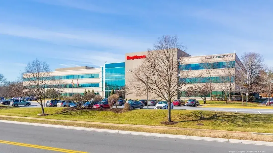 The Front of the office spaces at 5700 Rivertech with a parking lot of cars, a big company logo in red letters, and large glass windows.
