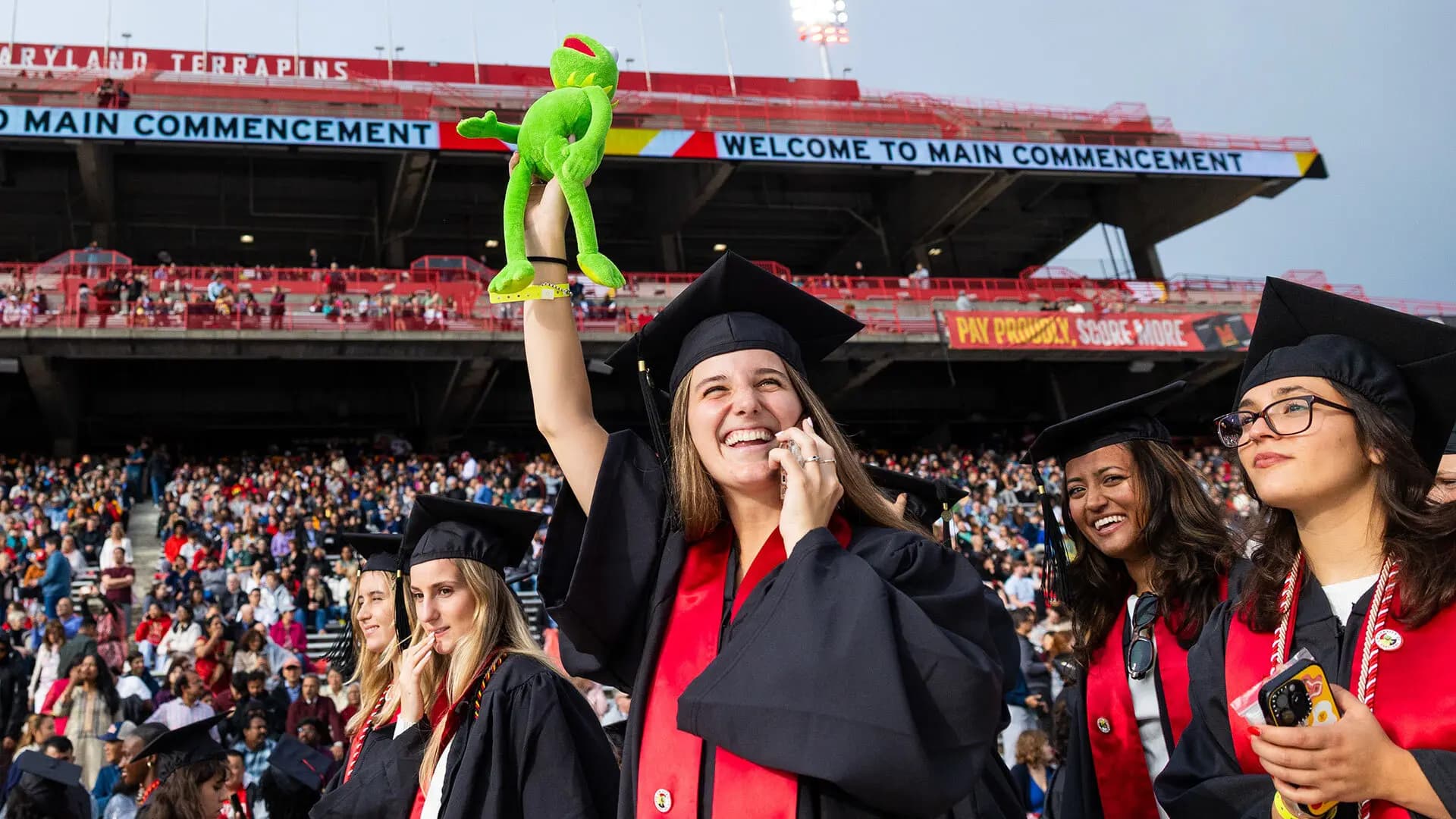 Graduate holding phone and stuffed Kermit, waving to crowd at SECU Stadium