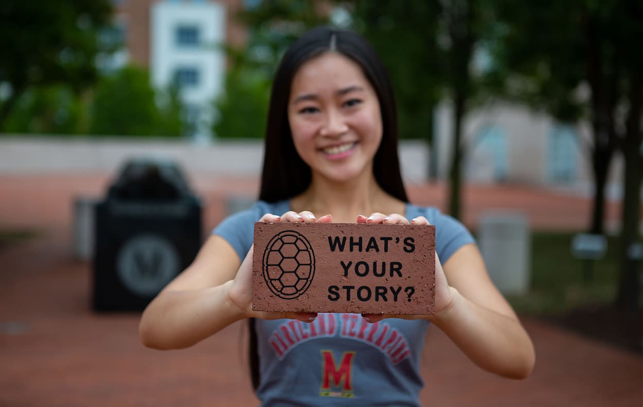 Image of graduate holding a brick engraved with the phrase, "What's Your Story?"