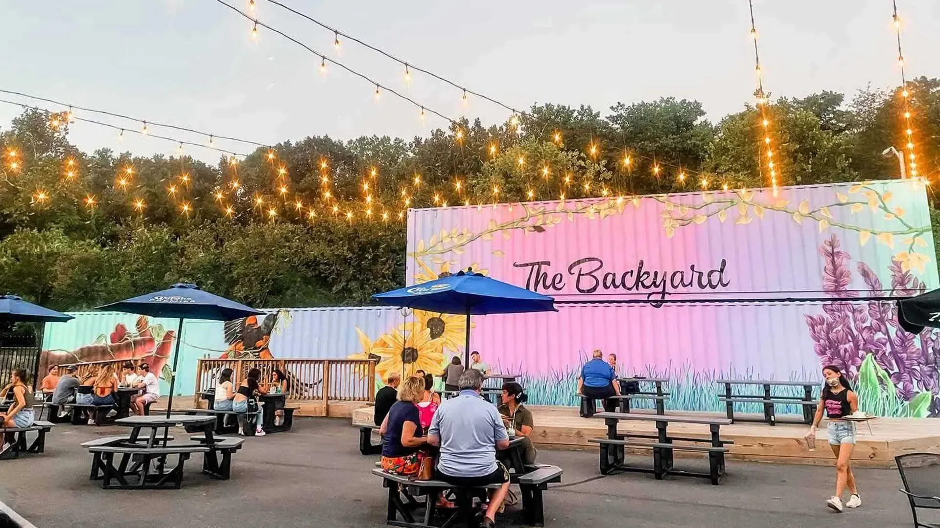 Outdoor tables with blue umbrellas and seated customers are scattered around a cement courtyard with a pink and purple mural on the wall behind the seating area and the words "The Backyard" painted in black.